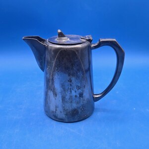 May include: A silver-colored metal coffee pot with a handle and a lid. The pot has a spout for pouring and a curved handle. The lid has a small handle on top. The pot is set against a solid blue background.