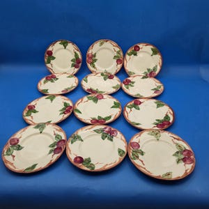 May include: A set of eleven round ceramic plates featuring a cream-colored base with hand-painted illustrations of purple plums and green leaves along the edges. Each plate has a thin brown rim and a smooth, glossy finish on the surface.