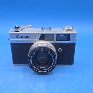 May include: A vintage Canon camera with a silver top and black leather-like body. The lens is prominent, with the text "CANON LENS SE 45mm 1:1.9" visible. The camera also has the text "Canon QUICK LOADING" on the front.