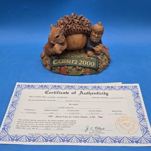May include: A Cairn Studio figurine featuring a squirrel and a gnome sitting beside a large acorn, with a banner reading CAIRN 2000. Included is a white Certificate of Authenticity with blue decorative borders and printed text detailing the artwork.