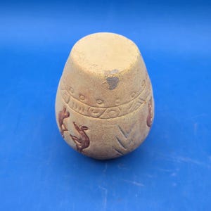 A view of the light brown terracotta vase from the bottom, showing its flat base and the etched Kokopelli figures and tribal patterns on its curved side.