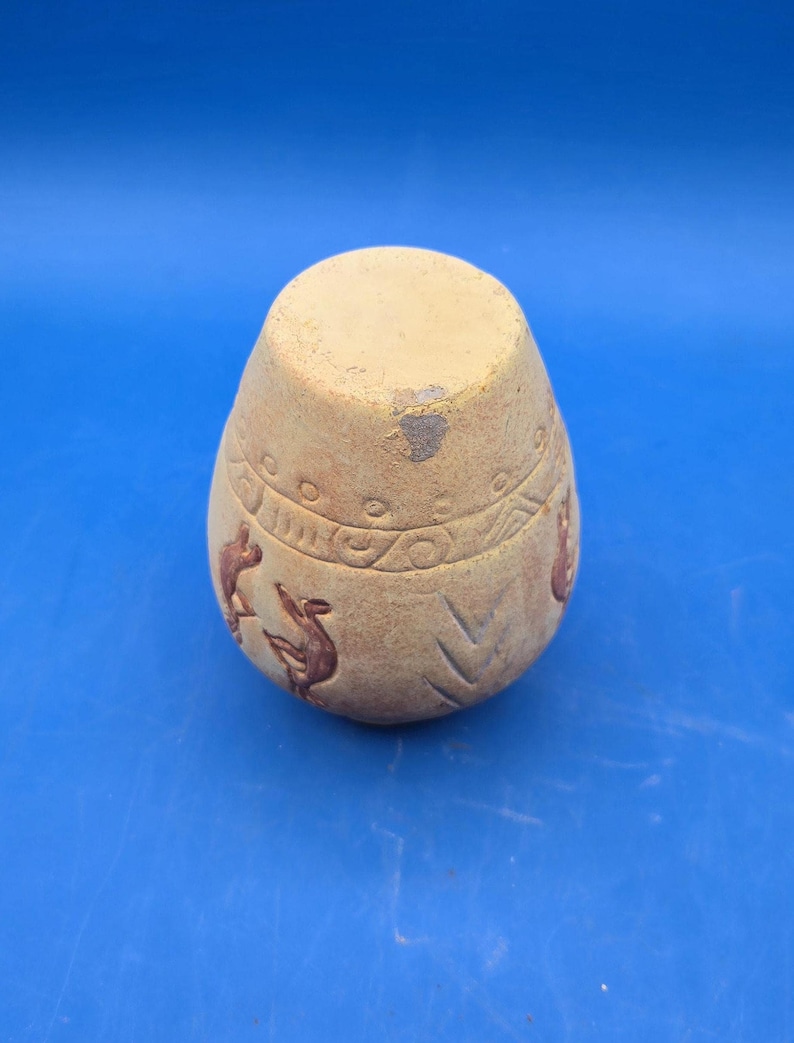 A view of the light brown terracotta vase from the bottom, showing its flat base and the etched Kokopelli figures and tribal patterns on its curved side.