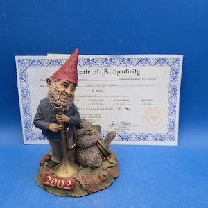 May include: A Cairn Studio figurine of a gnome with a red hat and blue jacket playing a gold trumpet, accompanied by a small brown animal playing a horn. The base features the text 2002 and Music to the Years, accompanied by a Certificate of Authenticity.