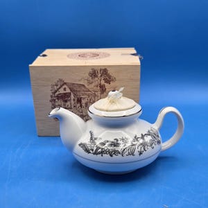 May include: White ceramic teapot with black floral design and gold trim. The teapot has a curved handle and spout, and a lid with a decorative top. A wooden box with a cabin illustration is in the background.