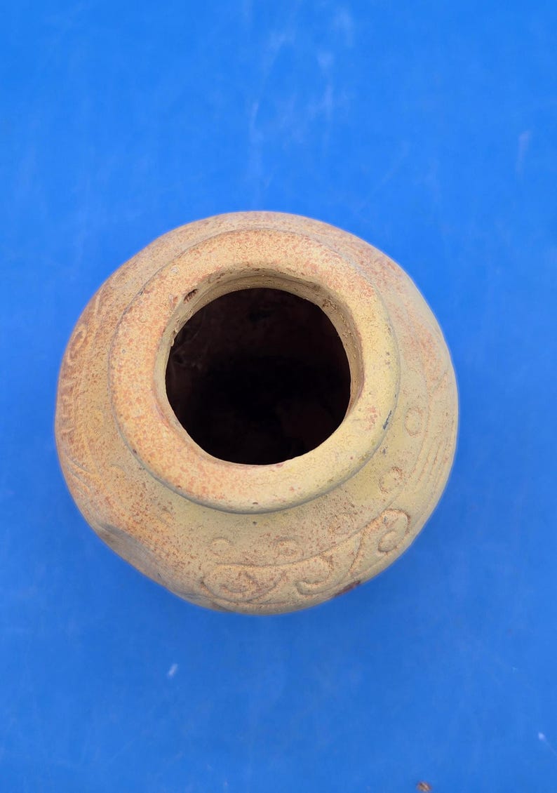 An overhead view of the light brown terracotta vase, showing the circular opening of its narrow neck and etched tribal patterns on the rim.
