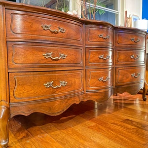 May include: A wooden chest of drawers with nine drawers and decorative brass handles. The chest has a curved front and stands on curved legs. The wood has a warm, reddish-brown tone, with drawers in three rows.