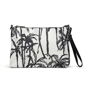 May include: A white clutch bag featuring a black palm tree design. It has a black zip closure and a black wrist strap. The pattern depicts a repeating motif of palm trees in a tropical forest.