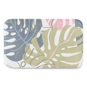 Puede incluir: Alfombra de baño rectangular con diseño botánico. Presenta grandes hojas de monstera estilizadas en tonos verdes, azules y rosas sobre un fondo blanco. El diseño tiene una estética moderna y artística.