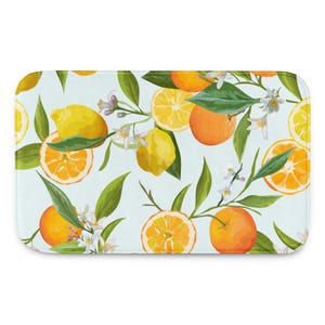 May include: A rectangular bath mat featuring a repeating pattern of bright yellow lemons, vibrant orange citrus slices, and small white blossoms with green leaves set against a light blue background. The design covers the entire surface area.