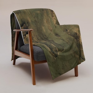 May include: A throw blanket featuring an abstract pattern of dark olive green, forest green, and earthy brown tones with textured, layered visual effects, draped over a dark gray armchair with a wooden frame and angled legs.
