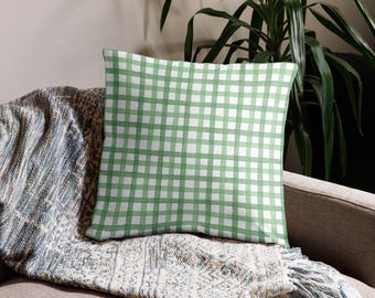 May include: A square throw pillow with a green and white gingham pattern. The pillow is resting on a gray sofa, with a blue and white patterned blanket underneath. A green plant is in the background.