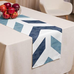 May include: A blue and white geometric patterned table runner. The runner features a repeating chevron design in shades of blue and white. The runner is placed on a beige tablecloth, with a gold wire basket of red apples in the background.