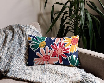 Puede incluir: Un cojín rectangular decorativo con un estampado floral sobre un fondo azul marino. Las flores son rojas, rosas, amarillas y verdes. El cojín está colocado sobre un sofá gris con una manta azul y blanca.