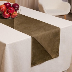 Puede incluir: Un camino de mesa marrón sobre un mantel blanco. Una cesta de alambre dorada con manzanas rojas sobre la mesa. Una silla blanca en el fondo.