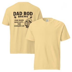 Puede incluir: Camiseta amarilla clara con el texto "DAD BOD BREWS" y un gráfico de jarra de cerveza. La parte trasera de la camiseta tiene el texto "FOR DADS WHO LIFT... THE COOLER LID". La camiseta está hecha de un material suave y cómodo.
