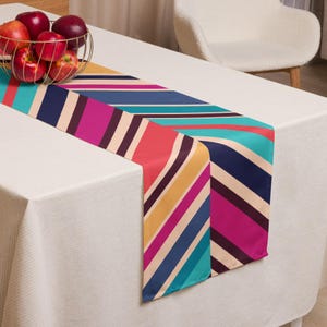 Può includere: Una colorata tovaglia con un motivo a righe diagonali nei toni del turchese, rosa, rosso, giallo e blu navy. La tovaglia è posizionata su una tovaglia bianca, con un cestino dorato di mele rosse sullo sfondo.