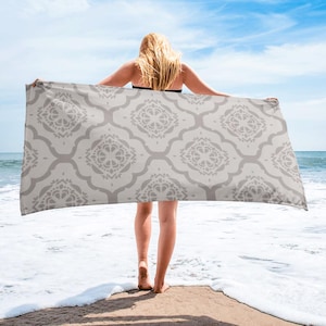 May include: A rectangular beach towel with a light gray and white geometric pattern. The towel is held up by a person standing on a sandy beach with the ocean and blue sky in the background.