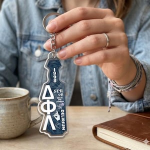 May include: A clear acrylic keychain with a silver chain. The keychain features white Greek letters and the text "1998 SPR 26 DELTA PHI LAMBDA DELIRIUM" in white. The background has a dark blue floral pattern.