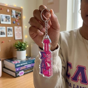 Puede incluir: Llavero acrílico transparente con un diseño rosa y morado. El llavero presenta el texto "1990", "ACE SPR 26", "ZETA SIGMA LAMBDA GAMMA" y "ROBIN". El diseño incluye rosas y una silueta de gato.