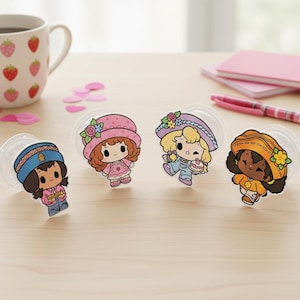 May include: Four colorful cartoon character pop sockets. Each character is a girl wearing a hat and outfit in different colors. The pop sockets are on a light wooden surface, with a strawberry-themed mug and pink stationery in the background.
