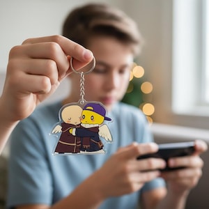 May include: A clear acrylic keychain featuring two cartoon figures hugging. One figure is bald and wearing a brown robe, the other is yellow with a purple cap and scarf. Both have white wings. The keychain is held up by a silver ring.