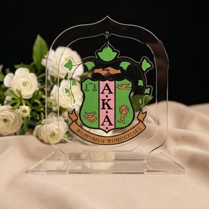 May include: Clear acrylic display featuring the Greek letters AKA in pink and black, set against a green shield with gold accents. The display includes the Greek phrase "ΦΕΛΩΜΕΝ ΨΗΦΗΤΙΔΕΣ" on a gold banner. White flowers are in the background.