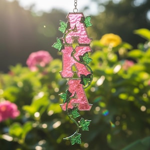 Puede incluir: Adorno colgante rosa y verde con las letras "AKA". Las letras están en una fuente decorativa y adornadas con hiedra verde. El fondo es una vista borrosa de un jardín con flores rosas y amarillas.