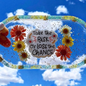 Könnte beinhalten: Ovale Resin-Tablett mit eingebetteten getrockneten Blumen in Rot-, Gelb- und Weißtönen. Das Tablett hat einen glitzernden Rand und enthält den Text "Take the Risk Or Lose the Chance".