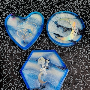 Puede incluir: Tres bandejas de resina en forma de corazón, redonda y hexagonal. Cada bandeja tiene un diseño veteado azul y blanco con detalles plateados. La bandeja en forma de corazón presenta un diseño de rosa y luna. La bandeja redonda tiene una luna y estrellas. La bandeja hexagonal tiene una rosa y una luna creciente.