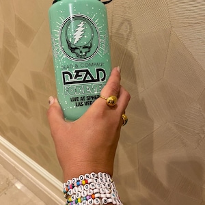 May include: A green water bottle with a white design featuring the Grateful Dead logo and the text "DEAD & COMPANY DEAD FOREVER LIVE AT SPHERE LAS VEGAS".