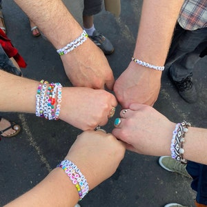 May include: Five people are wearing colorful beaded bracelets with different words spelled out on them. The bracelets are made of small plastic beads with letters on them. The people are holding their hands together in a fist bump.