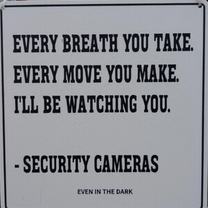 May include: White rectangular sign with black text. The sign reads: "EVERY BREATH YOU TAKE. EVERY MOVE YOU MAKE. I'LL BE WATCHING YOU. - SECURITY CAMERAS EVEN IN THE DARK."