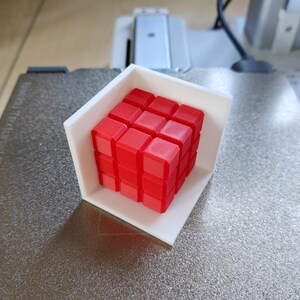 Pode incluir: Um cubo de Rubik vermelho 3x3, tamb&eacute;m conhecido como cubo m&aacute;gico, &eacute; exibido em uma moldura de pl&aacute;stico branca.