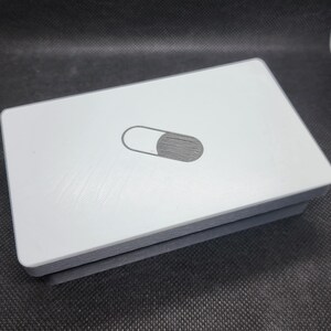 May include: A rectangular, light grey object featuring a dark grey oval design on its top surface. The object has a layered appearance with a darker grey base. The design resembles a switch or button.