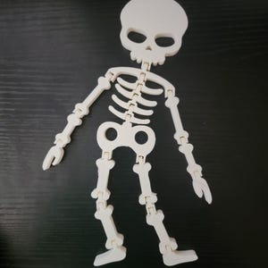 Puede incluir: Un juguete de esqueleto de plástico blanco con articulaciones. El esqueleto está en posición de pie con los brazos extendidos.