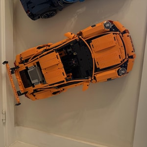 Könnte beinhalten: Ein orangefarbenes Lego-Modellauto mit schwarzen Innendetails. Das Auto ist an einer weißen Wand befestigt. Das Modell verfügt über detaillierte Merkmale, darunter Scheinwerfer und ein sichtbarer Motorraum. Ein weiteres blaues Lego-Auto ist über dem orangefarbenen Auto zu sehen.