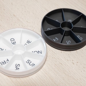 Puede incluir: Dos pastilleros redondos, uno blanco y otro negro, cada uno con siete compartimentos etiquetados para los días de la semana. El organizador blanco tiene una tapa translúcida, mientras que el negro es sólido.