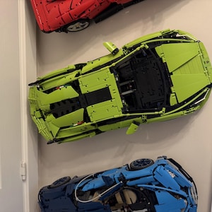 May include: Three toy sports cars in red, lime green, and blue are mounted on a wall. The cars are constructed from interlocking plastic bricks, showcasing detailed designs of the vehicles. The lime green car is in the center.