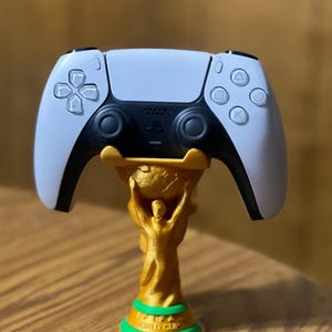 May include: A white and black PlayStation 5 controller resting on a gold-colored FIFA World Cup trophy stand with a green base. The controller has a symmetrical design with various button symbols.