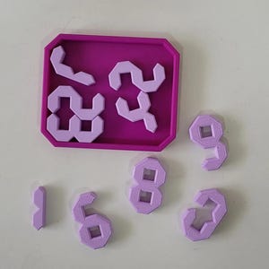 May include: A purple tray with a pink puzzle of the numbers 1, 2, 3, and 4. The puzzle pieces are arranged in a square shape. There are six additional pink puzzle pieces of the numbers 1, 2, 3, 4, 6, and 9.