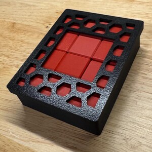 May include: A black box with a geometric pattern cut out of the top. The box contains a set of red square tiles.