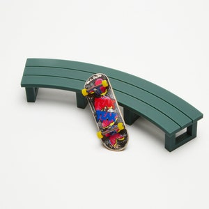 May include: A green plastic curved bench with a fingerboard skateboard on the ground in front of it.