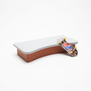 May include: A small, gray and red brick ramp with a curved edge. A fingerboard skateboard with a colorful graphic is lying on the ramp.