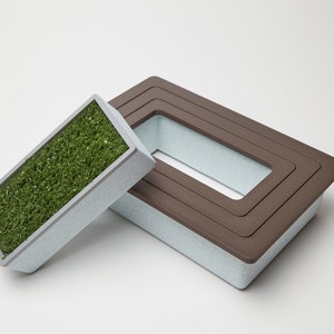 May include: A rectangular planter with a brown rim and a light gray base. The planter has a smaller rectangular opening inside. The planter is filled with green artificial grass.