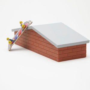 以下が含まれることがあります： 灰色の屋根と赤いレンガの土台を持つミニチュアレンガのランプ。黄色の車輪が付いたフィンガーボードスケートボードがランプの上に置かれています。