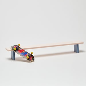 May include: A pink wooden ramp with blue supports, a fingerboard skateboard is resting on the ramp.