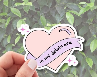 May include: Pink heart-shaped sticker with a purple ribbon that says "in my delulu era".