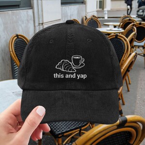 May include: Black corduroy baseball cap with a white embroidered design of a croissant and coffee cup, with the text "this and yap". The cap is being held up by a person's hand.