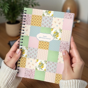 Puede incluir: Un diario con espiral con una cubierta de diseño patchwork. La cubierta presenta cuadrados de varias telas florales y estampadas en colores pastel, incluyendo rosa, amarillo y verde. El diario está etiquetado como "Lucy's Affirmation Journal".