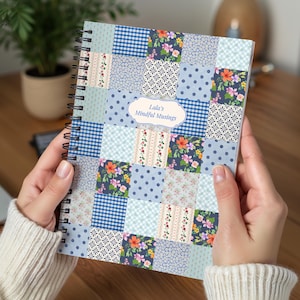 May include: A spiral-bound notebook with a patchwork design in shades of blue, featuring floral, polka dot, and gingham patterns. The cover includes the text "Lala's Mindful Musings" in a decorative label. The notebook is held by a person.